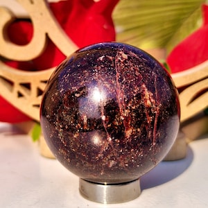 May include: A polished, dark red sphere with a speckled pattern, on a silver metal base. The sphere is set against red roses and a wooden decorative element.
