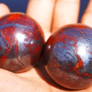 Op de afbeelding: Twee gladde, gepolijste bollen van rode en blauwe hematiet, elk met een uniek, draaiend patroon. De bollen zijn ongeveer zo groot als een golfbal.