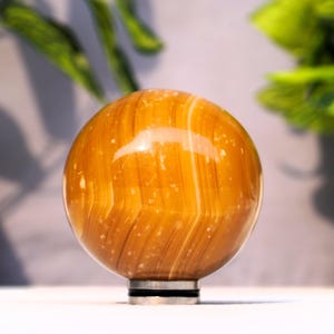 May include: A polished, golden-brown sphere with white striations. The sphere is made of natural stone and is displayed on a black stand.