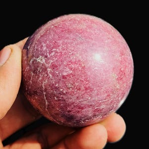 Op de afbeelding: Een gepolijste, bolvormige edelsteen in roze en magenta tinten, vastgehouden in een hand tegen een zwarte achtergrond. Het getextureerde oppervlak van de steen vertoont kleurvariaties, wat wijst op een natuurlijke minerale samenstelling. Diameter ongeveer 6 cm.