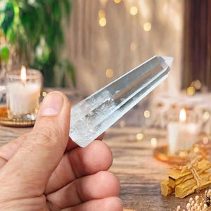 May include: A clear, faceted quartz crystal point with a hexagonal shape and a pointed tip. The crystal is transparent with visible internal inclusions and natural fractures, displaying a smooth, polished surface that reflects light.
