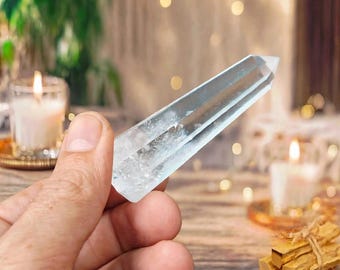 May include: A clear, faceted quartz crystal point with a hexagonal shape and a pointed tip. The crystal is transparent with visible internal inclusions and natural fractures, displaying a smooth, polished surface that reflects light.