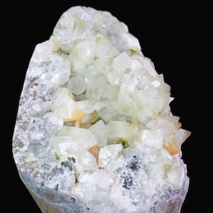 Puede incluir: Un espécimen de geoda natural con una estructura cristalina. La geoda presenta un grupo de cristales verde pálido y transparentes, con toques de naranja y una base gris-verdosa. El fondo es negro.