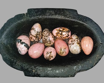 May include: A collection of nine pink and patterned gemstones, resembling eggs, nestled in a dark, textured, oval-shaped bowl. The gemstones vary in color, with some displaying black and brown veining.