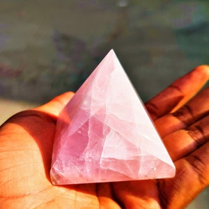 May include: A pink rose quartz pyramid crystal, a popular crystal for meditation and healing, is held in a person's hand.