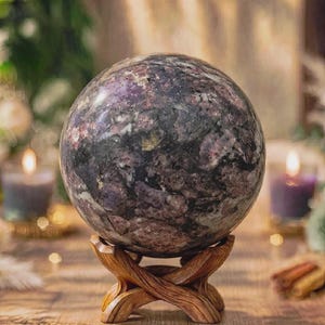Könnte beinhalten: Ein polierter, kugelförmiger Stein mit einer gesprenkelten Textur in tiefem Violett, Dunkelgrau und Schwarz. Die Kugel ruht auf einem geschnitzten, braunen Holzständer mit einem strukturierten, astähnlichen Design und vier geschwungenen Beinen.