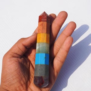 Könnte beinhalten: Ein sieben-Chakra-Kristallstab mit einer spitzen Spitze. Der Stab besteht aus sieben verschiedenen farbigen Steinen, die jeweils ein anderes Chakra repräsentieren. Die Steine sind rot, orange, gelb, grün, blau, indigo und violett.