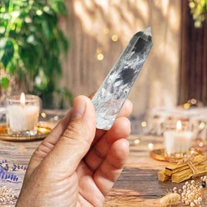 May include: A clear, faceted quartz crystal point with visible internal inclusions and fractures, featuring a hexagonal shape that tapers to a point at the top, held upright to display its translucent and reflective crystalline structure.
