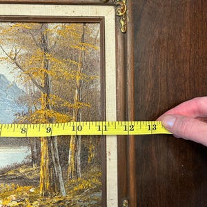 May include: A framed landscape painting with a brown wooden frame and gold corner accents. The painting depicts trees with yellow leaves, a body of water, and a mountain range. The painting measures approximately 13 inches in length.