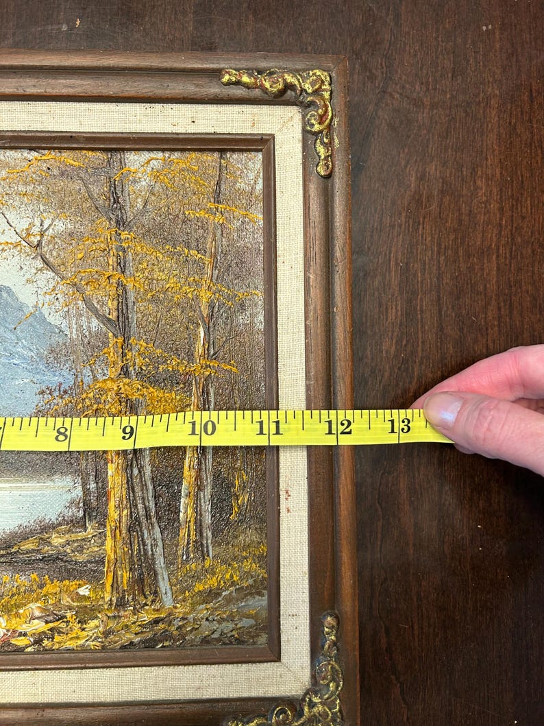 May include: A framed landscape painting with a brown wooden frame and gold corner accents. The painting depicts trees with yellow leaves, a body of water, and a mountain range. The painting measures approximately 13 inches in length.