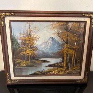 May include: Oil painting of a mountain landscape with a lake and trees. The painting has a dark brown wooden frame with gold corner accents. The trees have yellow leaves, and the mountain is blue.