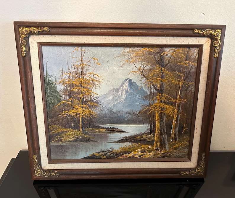 May include: Oil painting of a mountain landscape with a lake and trees. The painting has a dark brown wooden frame with gold corner accents. The trees have yellow leaves, and the mountain is blue.