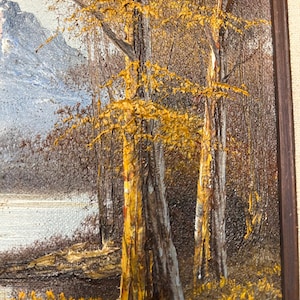 May include: Oil painting of a landscape featuring trees with golden yellow leaves and textured bark. A body of water and a mountain range are visible in the background. The artwork is framed in a dark brown wooden frame.