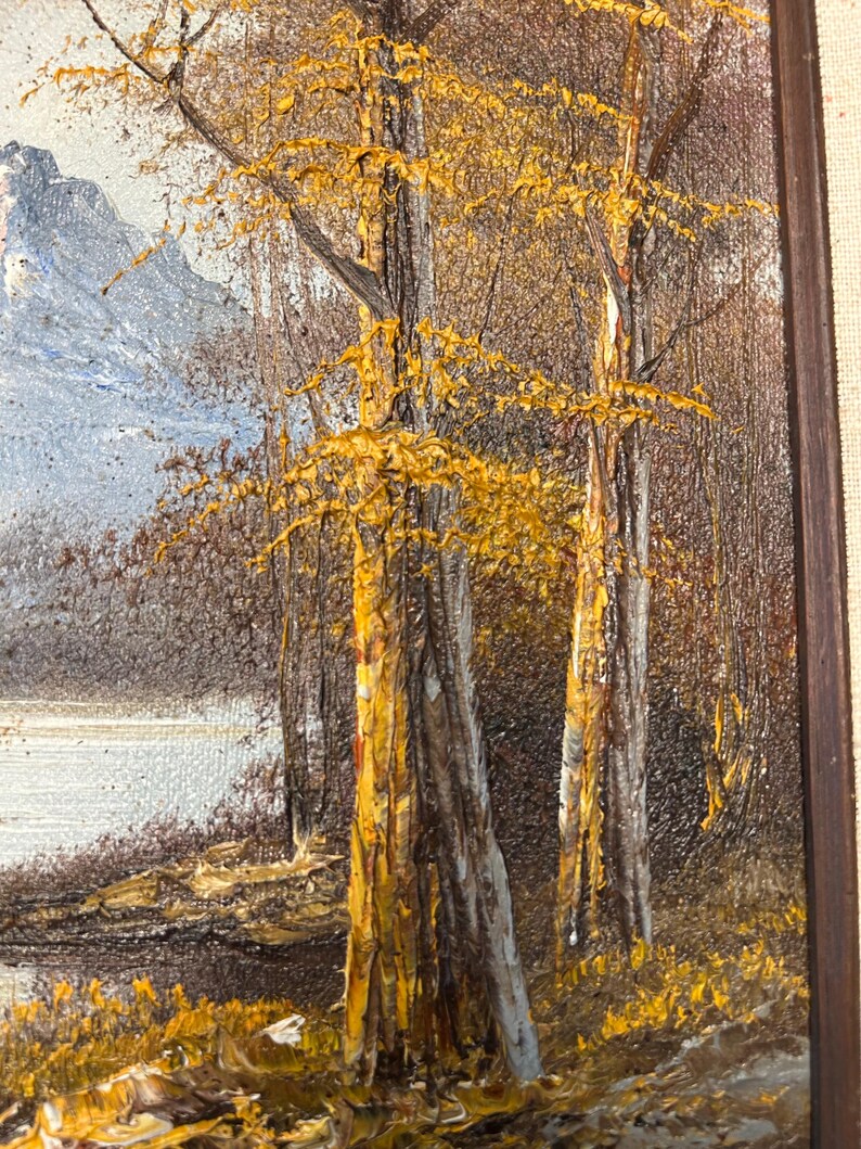 May include: Oil painting of a landscape featuring trees with golden yellow leaves and textured bark. A body of water and a mountain range are visible in the background. The artwork is framed in a dark brown wooden frame.
