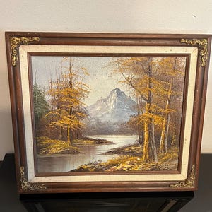 May include: Oil painting of a mountain landscape with a lake and trees. The painting features a mountain in the background, a lake in the middle, and trees with yellow leaves on either side. The painting is framed in a brown wooden frame with gold accents.