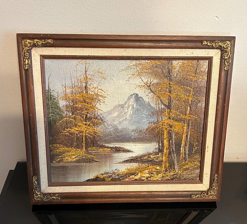 May include: Oil painting of a mountain landscape with a lake and trees. The painting features a mountain in the background, a lake in the middle, and trees with yellow leaves on either side. The painting is framed in a brown wooden frame with gold accents.