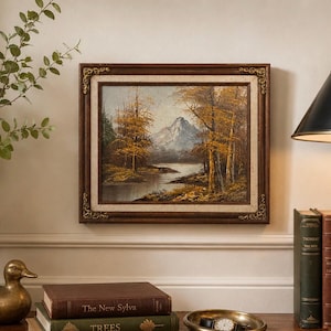 May include: An oil painting of a landscape scene with a mountain, trees, and a river. The painting is in a dark brown ornate frame. The artwork is displayed on a cream-colored wall, with books and a lamp nearby.