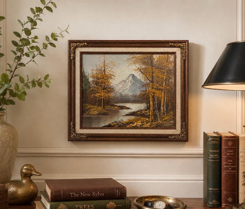 May include: An oil painting of a landscape scene with a mountain, trees, and a river. The painting is in a dark brown ornate frame. The artwork is displayed on a cream-colored wall, with books and a lamp nearby.