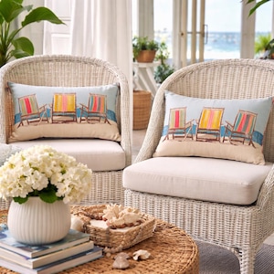 May include: Two white wicker armchairs with beige cushions. Each chair has a rectangular pillow with a beach scene featuring colorful beach chairs. A round, woven coffee table holds a white vase of flowers and a woven basket with seashells.