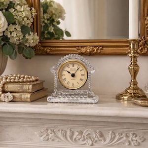 May include: A clear crystal quartz clock with a gold-colored face and black Roman numerals. The clock is set in a decorative crystal frame. The clock is on a white surface with a gold-framed mirror in the background.