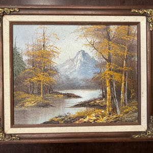 May include: Oil painting of a landscape scene with a lake, trees, and a mountain in the background. The trees have yellow leaves, and the painting is framed in a dark wood frame with gold corner accents.
