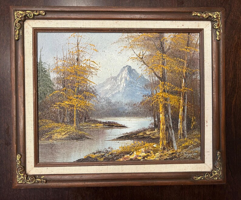 May include: Oil painting of a landscape scene with a lake, trees, and a mountain in the background. The trees have yellow leaves, and the painting is framed in a dark wood frame with gold corner accents.