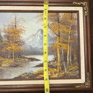 May include: Oil painting of a landscape with a river, trees, and a mountain in the background. The painting is in a brown wooden frame, and the image is approximately 10 inches tall.