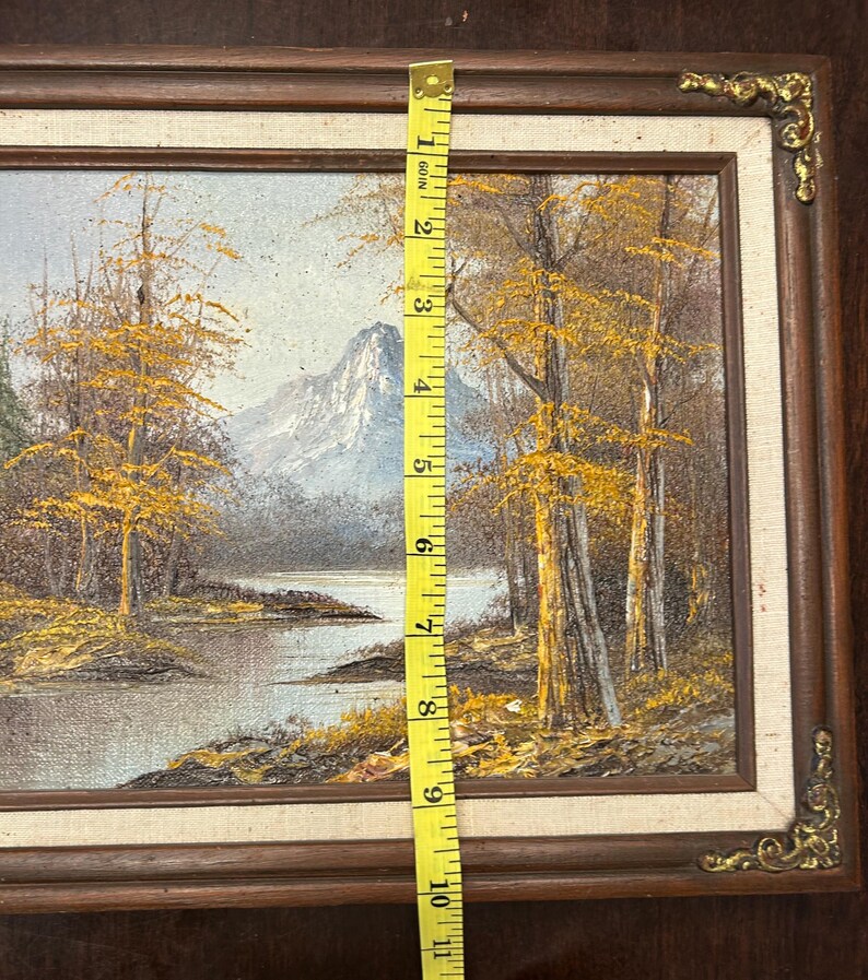 May include: Oil painting of a landscape with a river, trees, and a mountain in the background. The painting is in a brown wooden frame, and the image is approximately 10 inches tall.