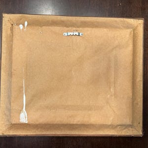 May include: Back of a framed artwork. The frame is brown, and the backing is a light brown paper. A metal sawtooth hanger is attached to the top center. White paint splatters are visible on the backing.