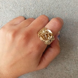 Könnte beinhalten: Ein goldfarbener Ring mit einem detaillierten Rosenmotiv. Der Ring wird an einem Finger getragen.