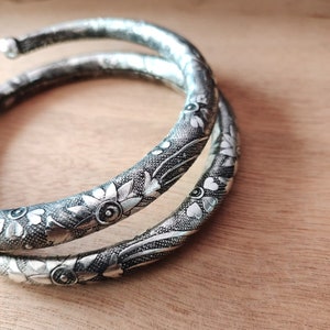 Puede incluir: Dos brazaletes de plata con intrincados diseños florales. Los brazaletes están apilados uno encima del otro y tienen un acabado brillante.