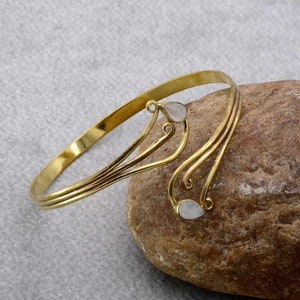 Könnte beinhalten: Ein goldfarbenes Armband mit einem zarten, fließenden Design. Das Armband hat zwei weiße, tropfenförmige Steine.