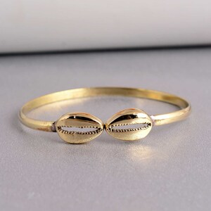 Könnte beinhalten: Ein goldenes Armreif-Armband mit zwei muschelförmigen Anhängern. Das Armband ist schlicht und elegant.