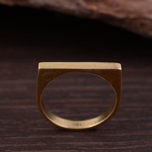 Könnte beinhalten: Ein goldfarbener Ring mit rechteckiger Oberseite und abgerundetem Band. Der Ring besteht aus massivem Metall und hat ein schlichtes, modernes Design. Der Ring wird auf einer dunklen Holzoberfläche gezeigt.