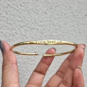 Könnte beinhalten: Goldfarbenes Armreif-Armband mit dem eingravierten Schriftzug "FUCK SHIT UP" auf der Vorderseite. Das Armband wird vor einem hellgrauen Hintergrund gehalten.