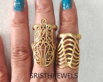 May include: Two gold rings are shown on fingers with blue nail polish. One ring has a Hamsa hand design, the other a cage-like structure. The rings are likely part of a jewellery collection. The text "SRISTHIJEWELS" is visible.