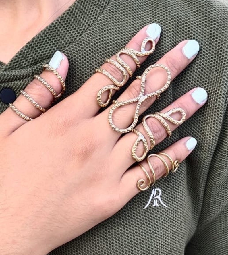 May include: A set of five gold-toned rings, featuring a variety of intricate designs. The rings are worn on the fingers of a hand, with some rings worn on multiple fingers.