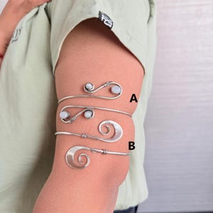 Könnte beinhalten: Drei silberne Armreifen mit weißen Steinen. Die Armreifen sind übereinander auf dem Arm einer Person gelegt. Der obere Armreif ist mit "A" und der untere mit "B" gekennzeichnet.