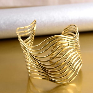 Könnte beinhalten: Ein goldfarbenes Metallarmband mit einem wellenförmigen, geschichteten Design. Das Armband ist offen und hat eine breite, abgerundete Form.
