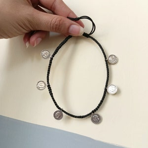 Puede incluir: Un brazalete de cordón negro con dijes de monedas plateadas. Las monedas tienen un perfil en relieve de una cara.