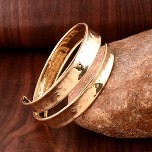 May include: Two gold-toned hammered metal bangles, one slightly larger than the other, are stacked on top of each other. The bangles are smooth and have a slightly textured surface.