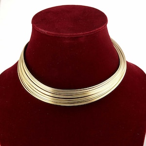 Könnte beinhalten: Eine goldfarbene Halskette mit mehreren Strängen, präsentiert auf einer roten Samtbüste. Die Halskette im Choker-Stil hat viele dünne, parallele Stränge, die einen kühnen, modernen Look erzeugen. Der Schmuck ist vor einem kontrastierenden Hintergrund platziert.