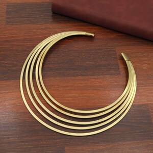 Könnte beinhalten: Eine goldfarbene Metallhalskette, bestehend aus fünf konzentrischen, halbkreisförmigen Bändern, die eine breite, offene Halbmondform bilden. Die glatten, polierten Metallbänder sind an den Enden verbunden und erzeugen ein strukturiertes Design.