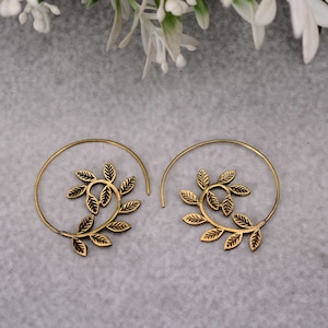 Puede incluir: Un par de pendientes en espiral de color dorado. Cada pendiente presenta un diseño de hojas en el centro de la espiral. Las hojas están detalladas con venas y están dispuestas en un patrón circular. Los pendientes están sobre un fondo neutro.