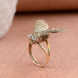 May include: A gold-toned ring featuring a detailed bee design with clear crystal accents for the eyes. The bee is positioned with its wings spread out.