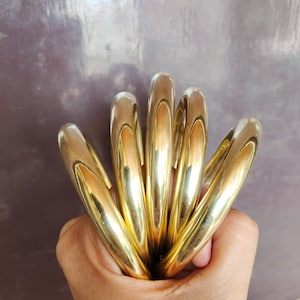Könnte beinhalten: Eine Sammlung goldfarbener Armreifen, die zusammengehalten werden. Die Armreifen sind glatt und glänzend und reflektieren das Licht. Der Schmuck hat eine warme, goldene Farbe, die auf einen klassischen oder Vintage-Stil hindeutet. Die Armreifen sind gestapelt und erzeugen einen auffälligen Look.