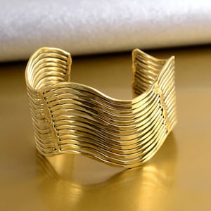Könnte beinhalten: Ein goldfarbenes Metall-Armband mit einem wellenförmigen, geschichteten Design. Das Armband besteht aus mehreren dünnen Metallstreifen, die miteinander verwoben sind, um einen einzigartigen und stilvollen Look zu kreieren.