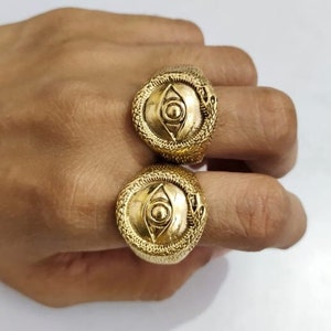 Könnte beinhalten: Zwei goldene Ringe mit einem Schlangendesign und einem Auge in der Mitte. Die Ringe werden an zwei Fingern einer Hand getragen.