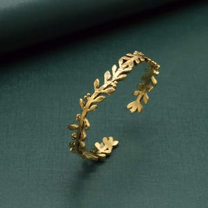 Könnte beinhalten: Ein goldfarbenes, offenes Armband mit Blattdesign. Das Armband hat detaillierte Blätter und kleine Beeren, die eine von der Natur inspirierte Ästhetik schaffen. Das Armband besteht aus glänzendem Metall.