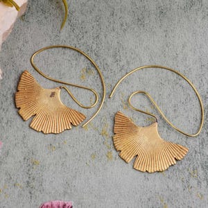Könnte beinhalten: Ein Paar goldfarbene Ohrringe. Jeder Ohrring hat ein fächerförmiges Design mit detaillierten Linien, das einem Ginkgoblatt ähnelt. Die Ohrringe haben einen gebogenen Draht, der durch das Ohr geht. Der Hintergrund ist grau und grün gefleckt.